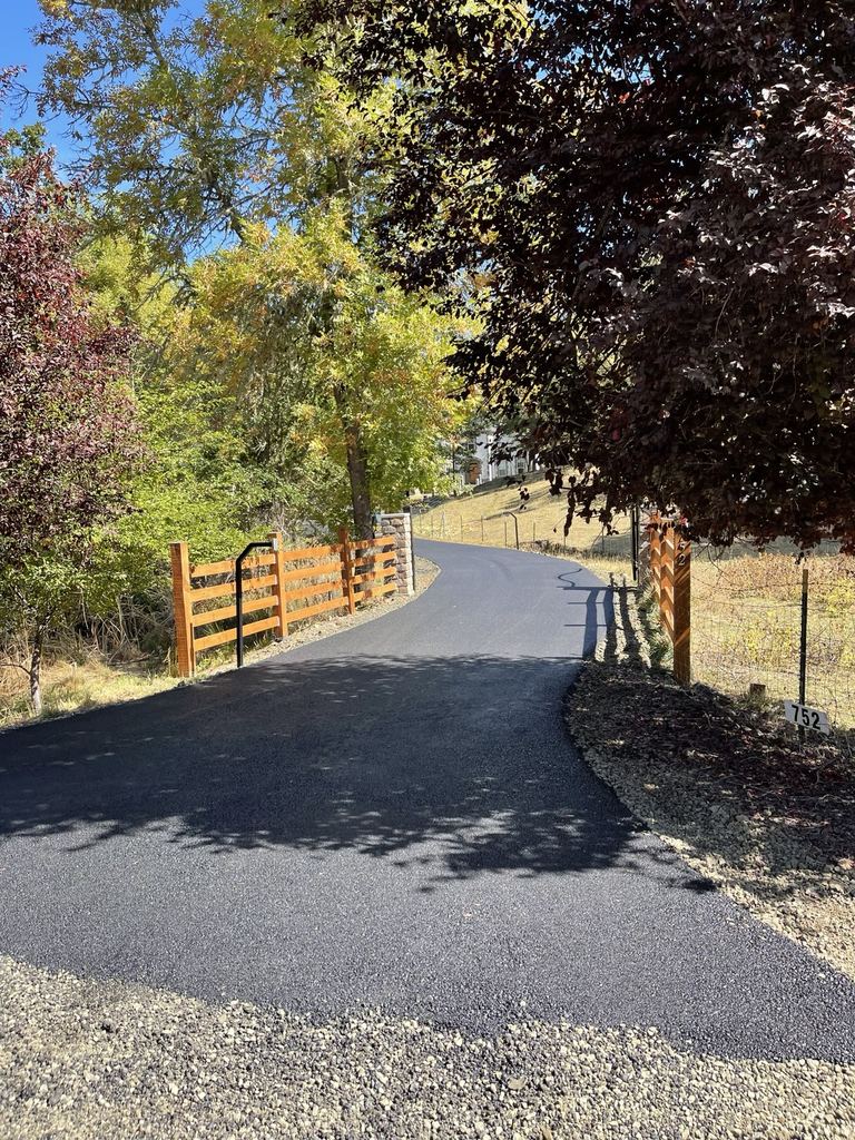 Asphalt drive passing between wood fencing and stone gateposts.