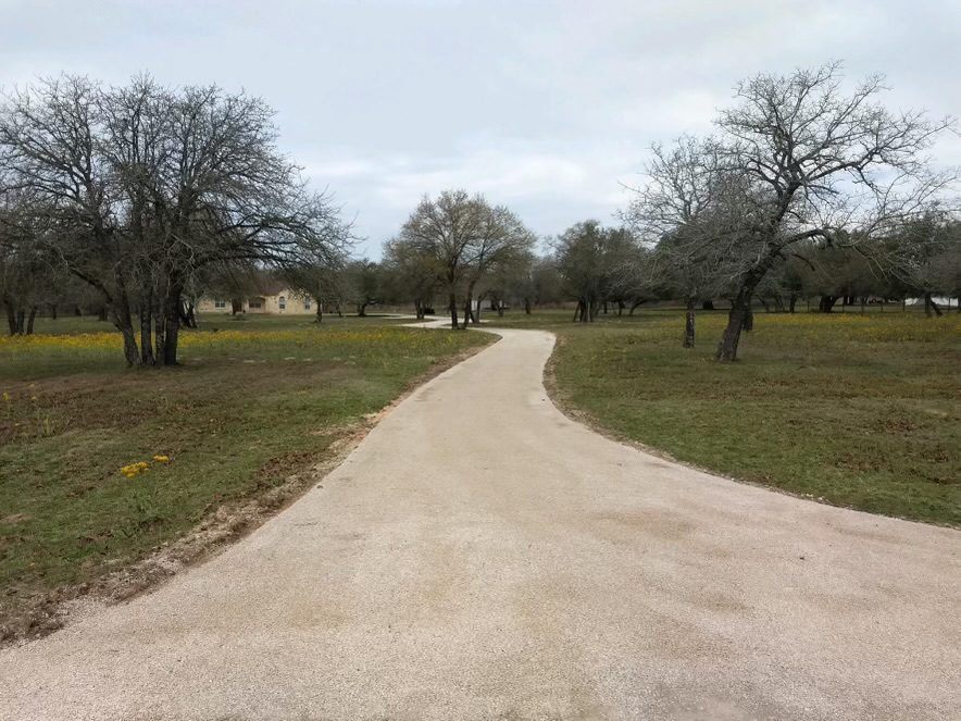 Long, pale chip seal drive curving through an open tree-lined property.