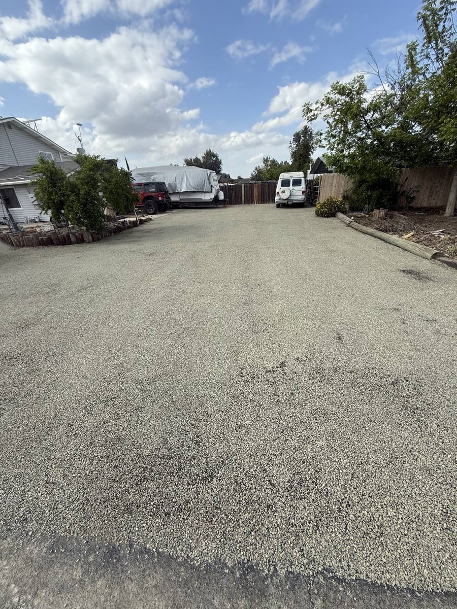 Wide chip seal parking area beside a home, with vehicles at the far end.
