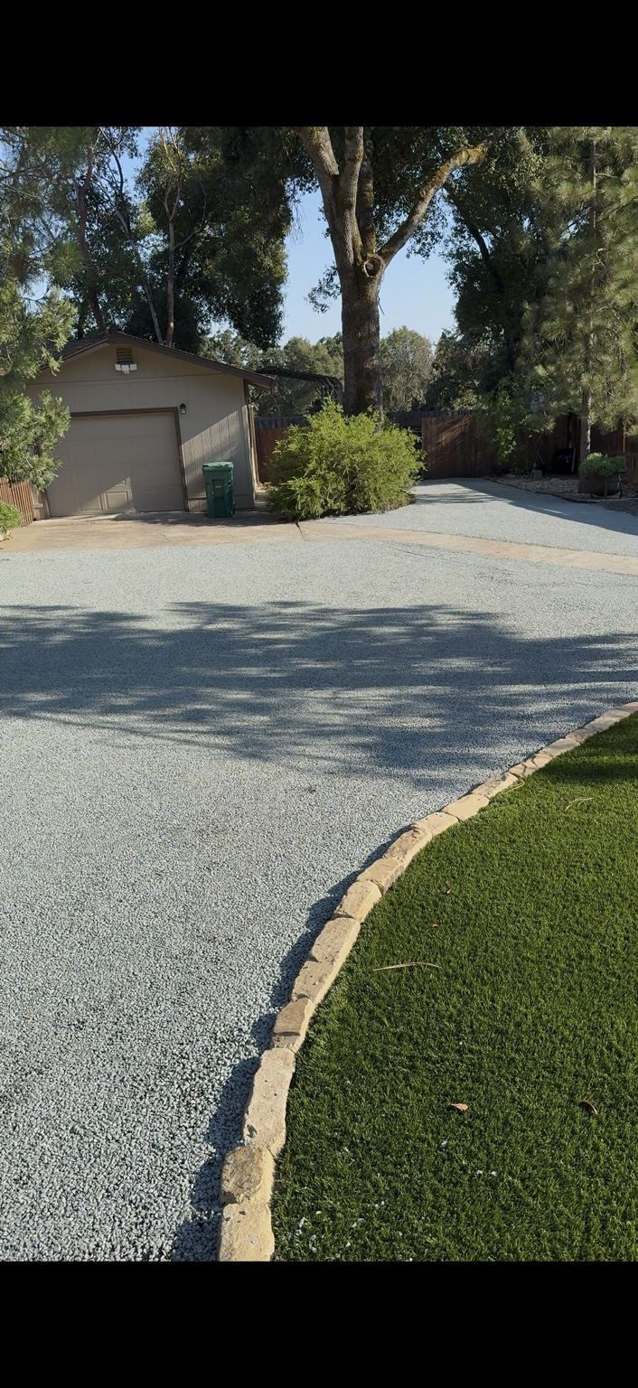 Gray chip seal surface following a curved stone border beside a lawn.